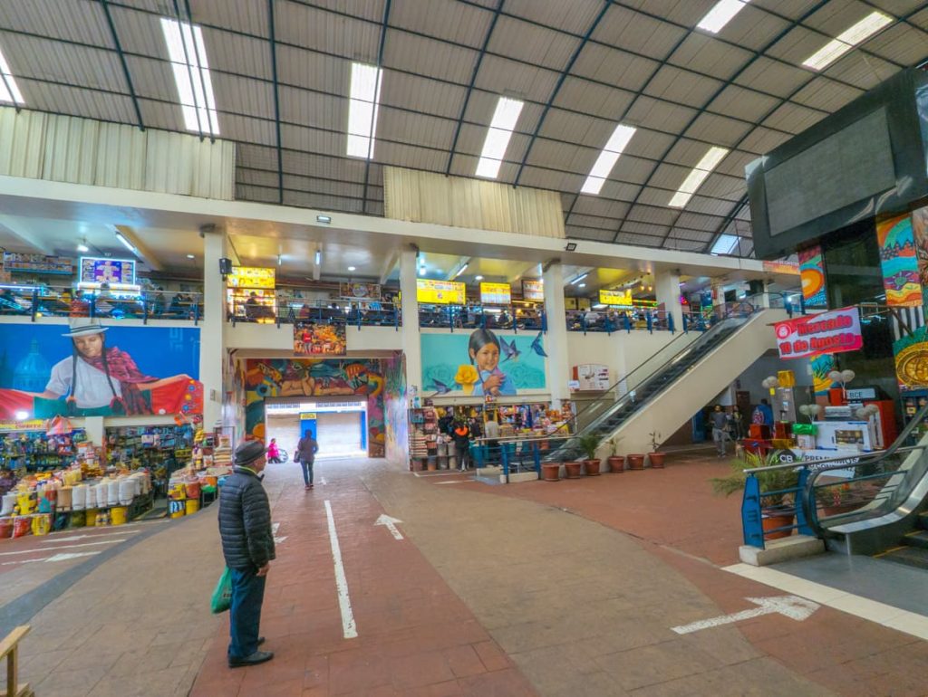 Interior of Mercado 10 de Agosto in Cuenca Ecuador, with food stalls, murals, and escalators connecting the upper and lower levels.