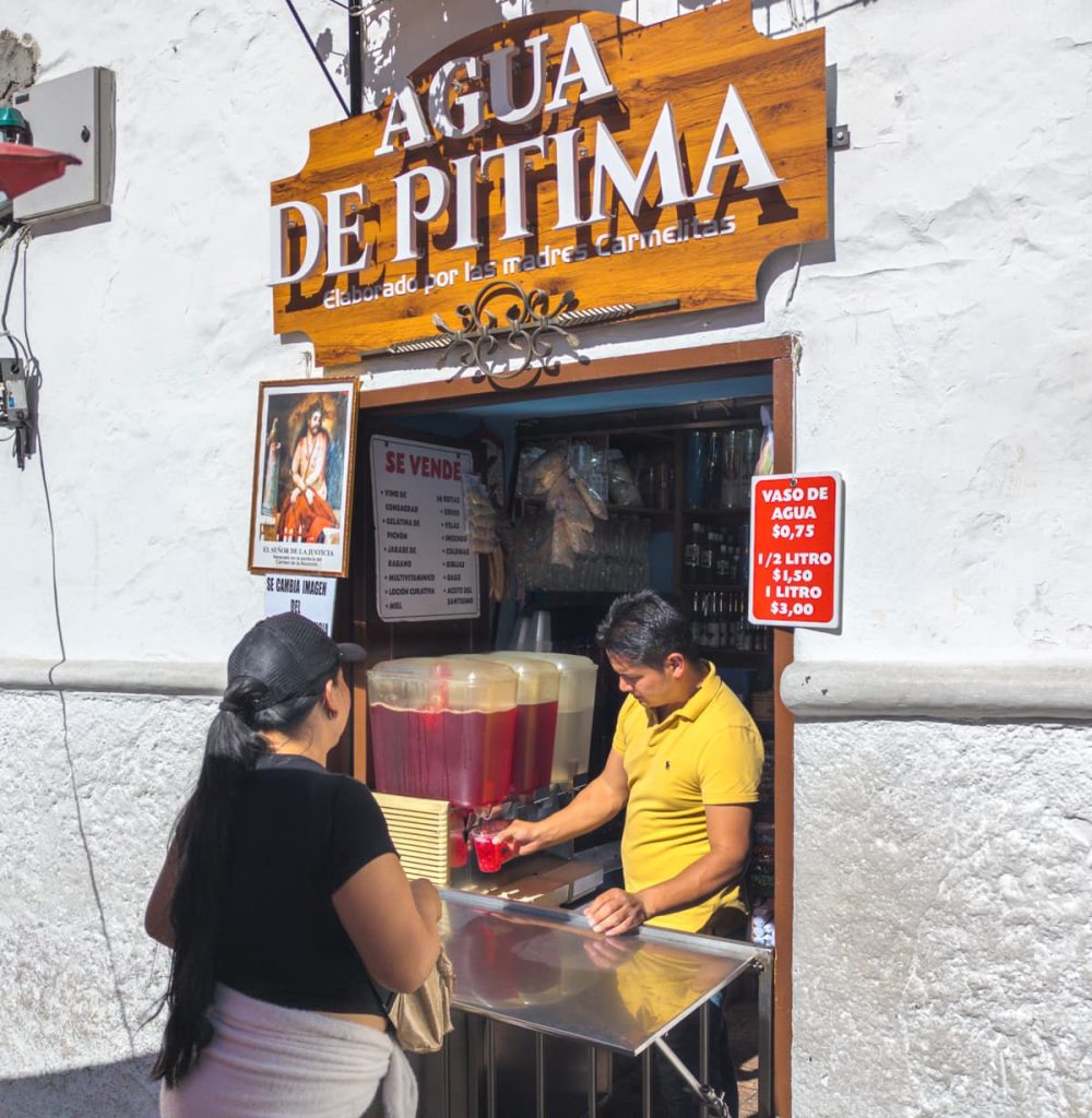 A street vendor in Cuenca serves a pink cup of agua de pitima, a traditional herbal drink made by nuns, to a customer from large drink dispensers. Trying this local refreshment is one of the unique things to do in Cuenca Ecuador.