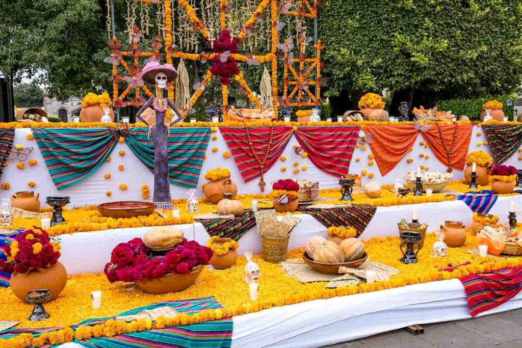 A white-clothed altar with different colorful cloths, lots of yellow flowers, pots, fruits, candles and a Day of the Dead statue.