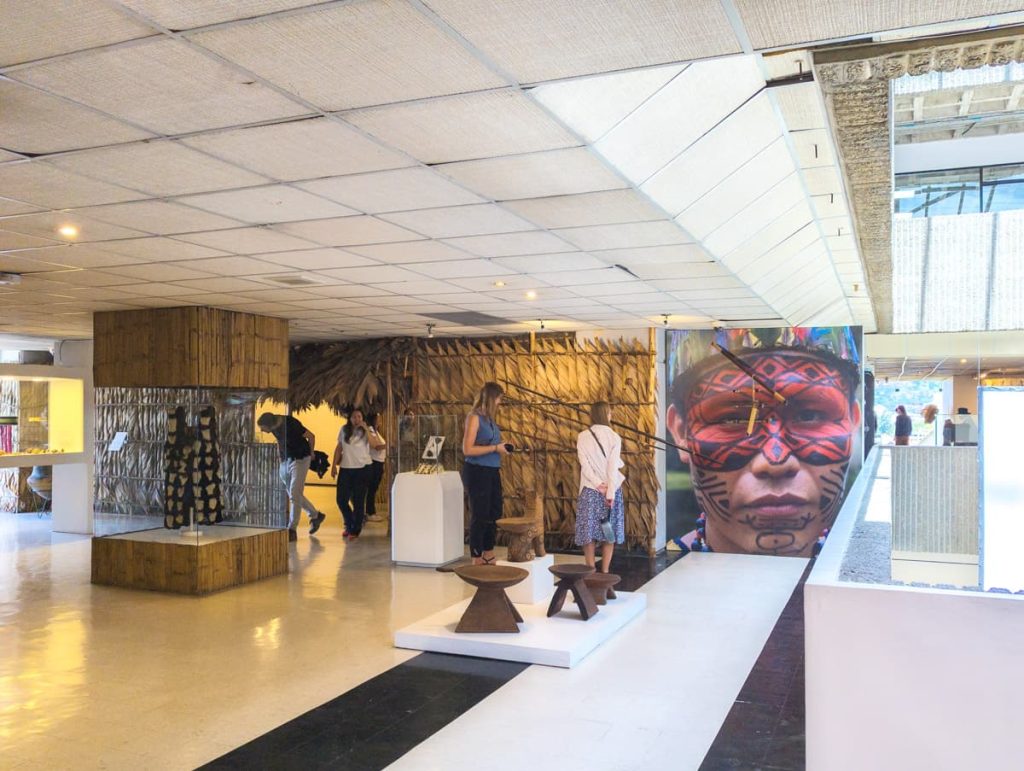 People wander among an interior exhibit at the Pumapungo Museum in Cuenca showcasing indigenous Amazonian culture, with traditional artifacts and a striking mural of a painted face.