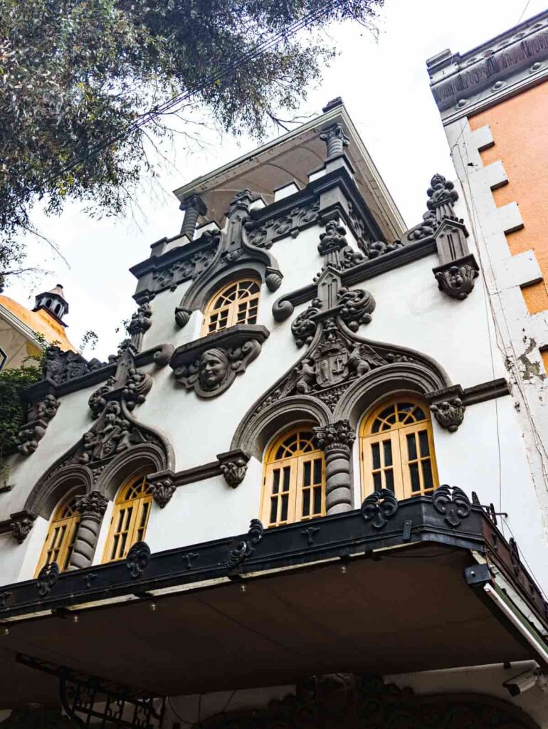 The ornate exterior of a building that showcases the architecture in Roma Norte Mexico City. A gargoyle face is mounted under the arched window in the center with other gargoyle structure decorating the other curved windows.