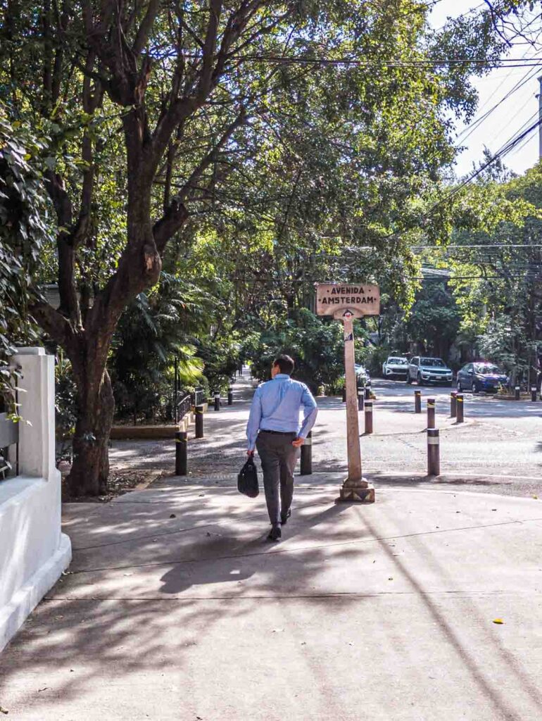 In La Condesa Hipódromo neighborhood in Mexico City, a man dressed in casual business attire walks away from the camera, passing an antique sign that says Avenida Amsterdam.