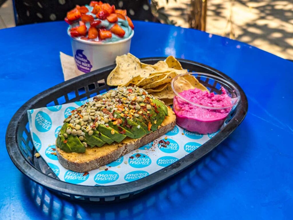 Sliced avocado on a piece of toast topped with a seed and nut mixture is served in a plastic basket with chips and a side of pink hummus at Mora Mora, a vegan restaurant in CDMX. In the background is an acai bowl topped with fresh strawberries.