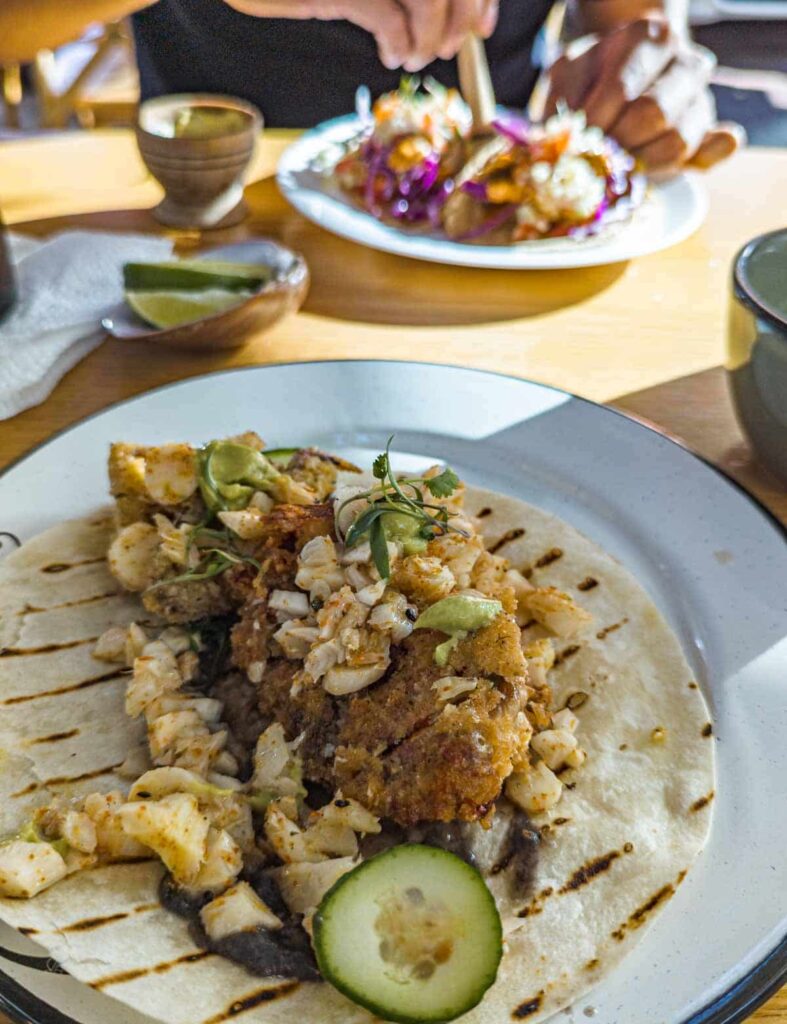 A white plate with a seafood taco topped with crab meat and cucumbers at Balandra Seafood in Mexico City. In the background, a second person squeezes lime on his fish tacos.