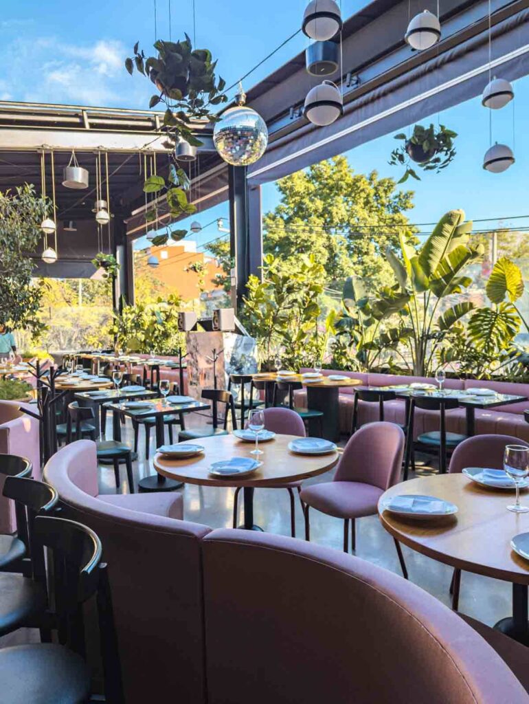 Pink couches and chairs at Balmori rooftop bar in Mexico City surrounded by plants. Above a disco ball hangs from the open roof which shows the blue sky.