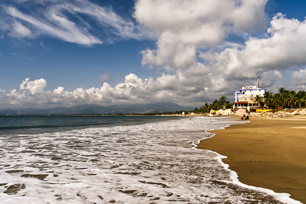 A shallow wave washes over the Barra de Potosi beach near Zihuatanejo Mexico. In the background are coconut palm trees, a white building, and mountains. The sky is full of white puffy clouds.