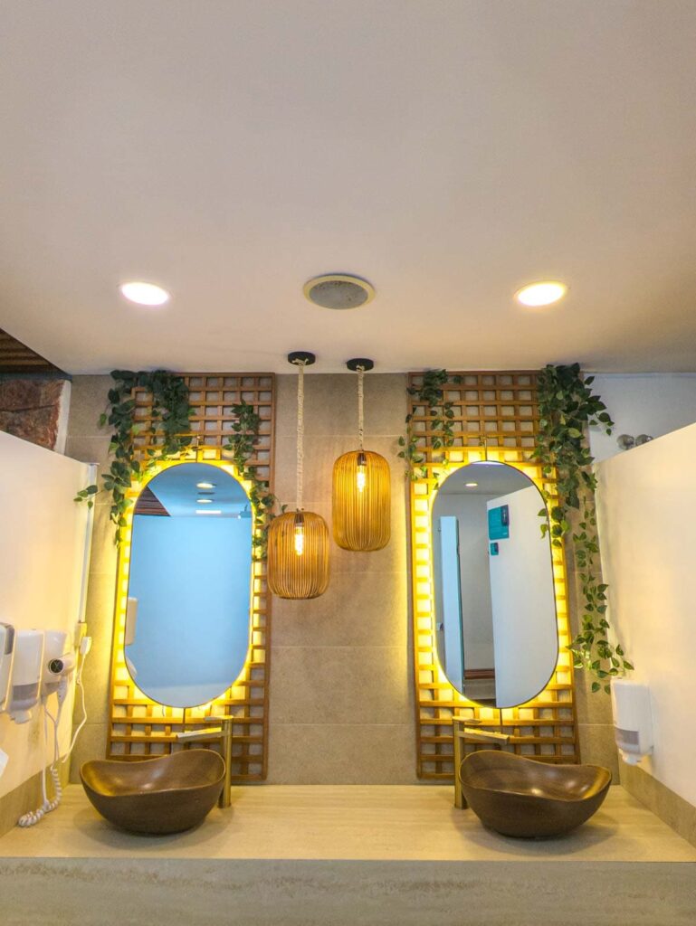 Stylish vanity area at Novaqua hot springs in Cuenca Ecuador, featuring two oval mirrors with warm backlighting, bronze vessel sinks, hanging bamboo lanterns, and climbing greenery.