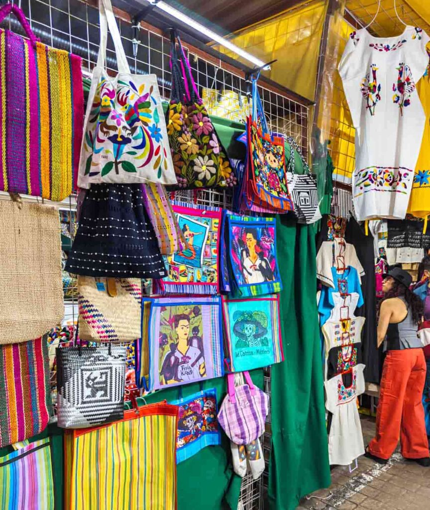 Colorful bags and other textiles hang at the artisan market in Coyoacán as a woman peeks inside a stand.