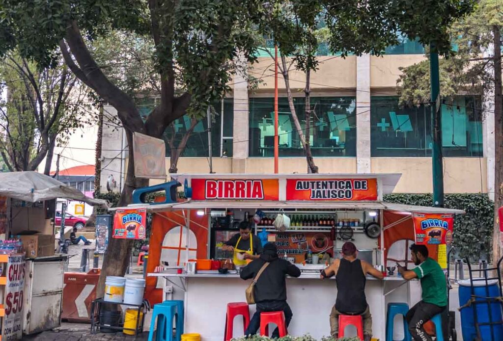 Several men sit around a taco stand advertising "autentica de Jalisco," birria tacos in Mexico City while the chef prepares tacos.