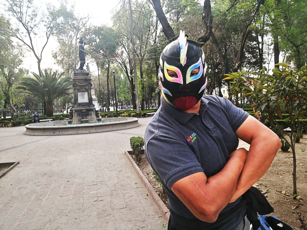During one of the best lucha libre tours in Mexico City, a former wrestler poses wearing a black luchador mask highlighted with rainbow accents and a mohawk. His arms are folded in front of him.