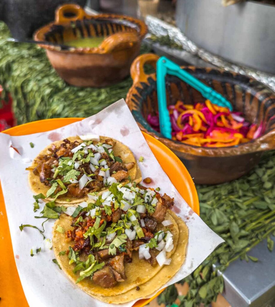 The best tacos in Mexico City on an orange plate and topped with cilantro and onion. In the background are large clay bowls. One is filled with pickled red onion and orange habanero peppers. The other is filled with a green salsa.