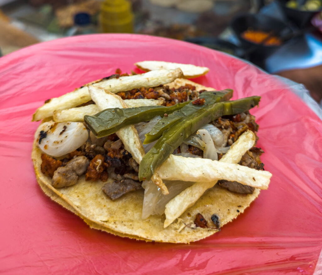A red plate, covered in a thin plastic bag holds a campechano taco topped with french fries, onion, and slices of nopal cactus.