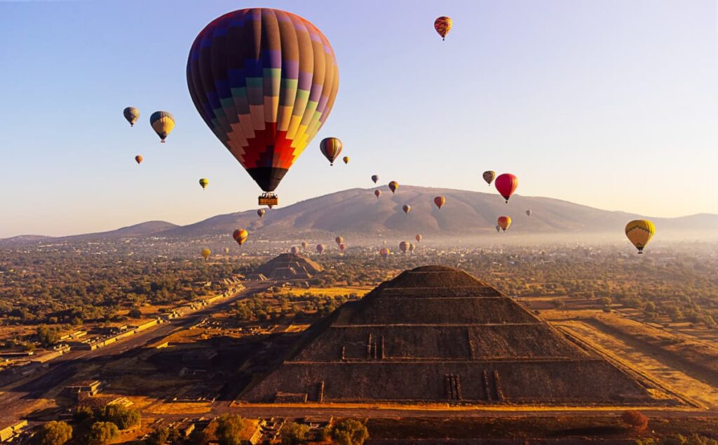 During a Teotihuacan balloon tour, more than a dozen colorful hot air balloons fly over the Teotihuacan pyramids. In the foreground is the Pyramid of the Sun, with the Pyramid of the Moon and mountains in the background.