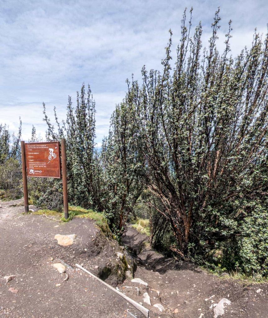 A dirt biking trail entrance marked by a brown signpost sits at the edge of a forested path on a mountain slope near the Teleferico in Quito.