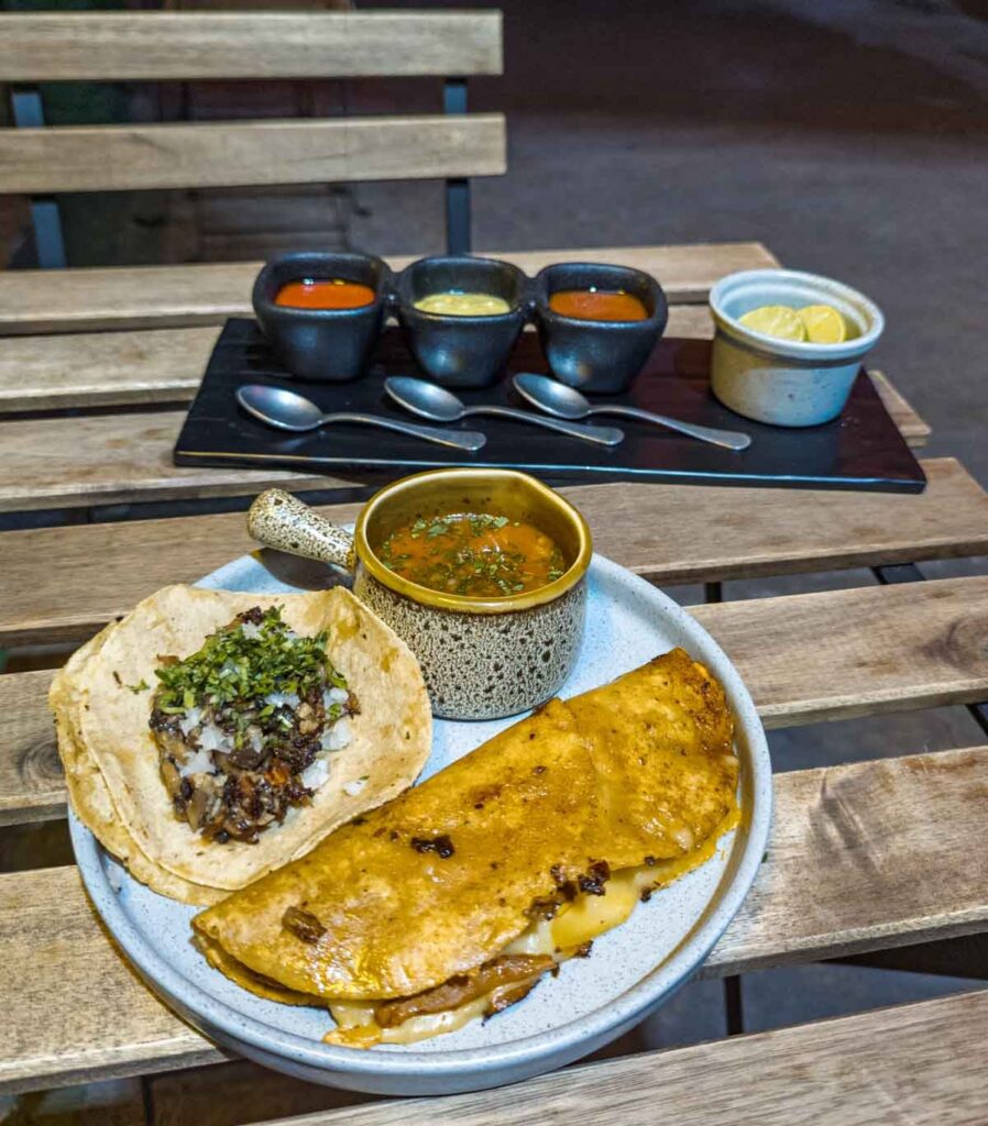 A vegan taco and quesabirria sit on a plate with a small bowl of consome at Alguito Vegana in Mexico City. In the background is a long plate with three bowls of different salsas and a bowl of limes.