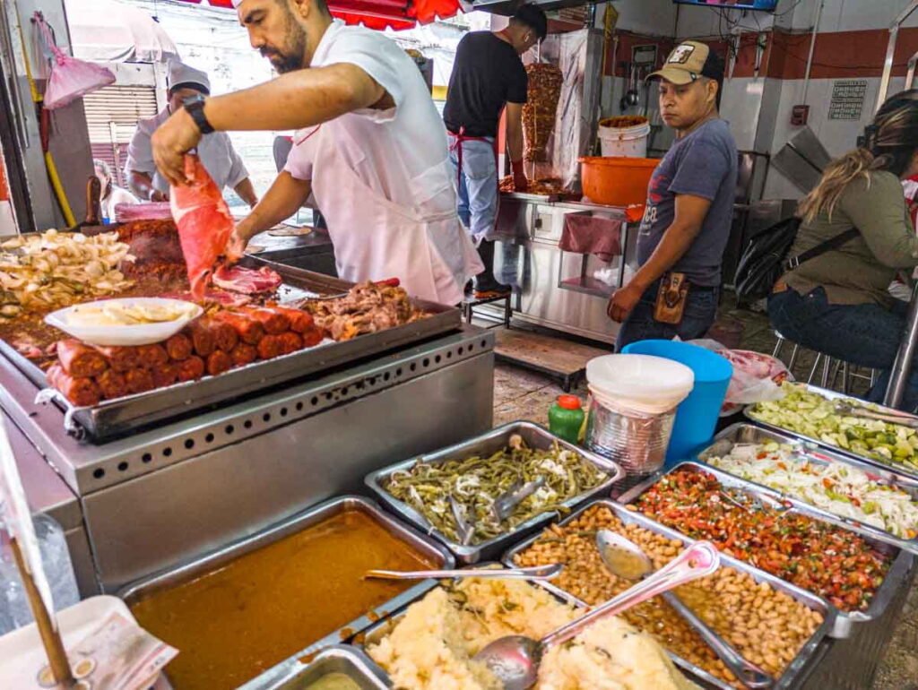 A man places a piece of bistek (thin beef) on the frill which is stacked with other meats such as chorizo. In the background a man layers meat onto a spit for tacos al pastor. And in the foreground are large rectangular pans of toppings such as mashed potatoes and pinto beans.