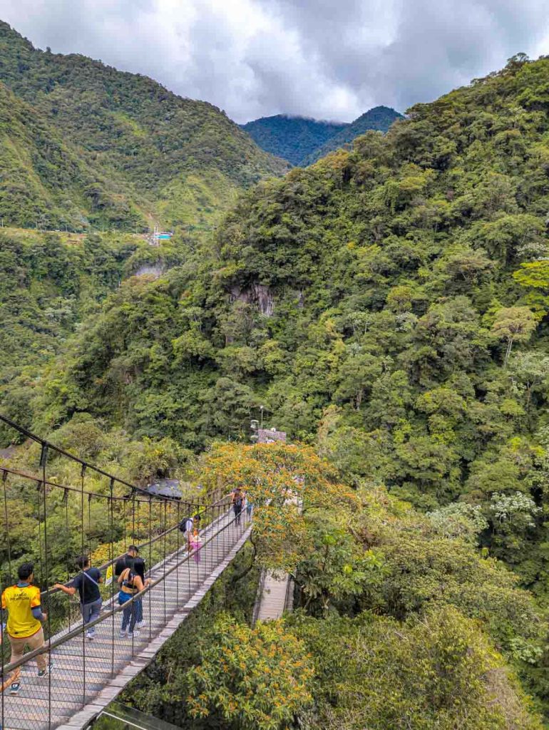 People walk across a suspension bridge high above the jungle canopy with views of green mountains at the Diablo waterfall in Baños, Ecuador.