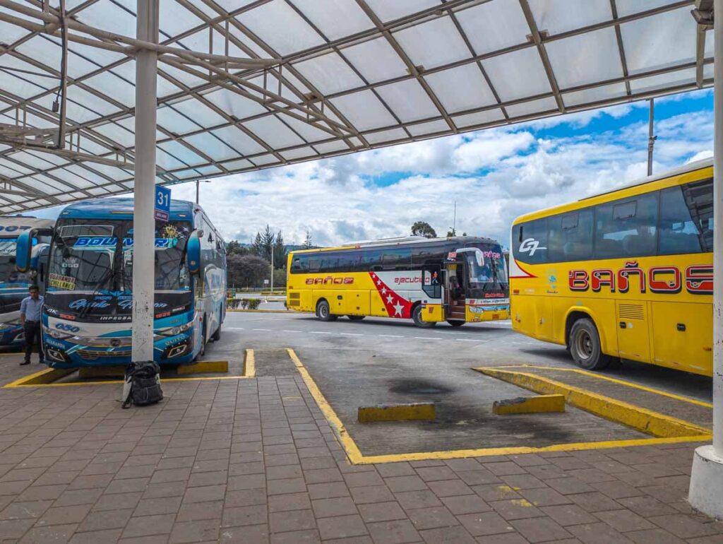 At the Quitumbe bus station in Quito, two buses are parked in the boarding area while another drives behind them. Two of them are the Quito to Baños bus - yellow with the name Baños in red lettering on the side.