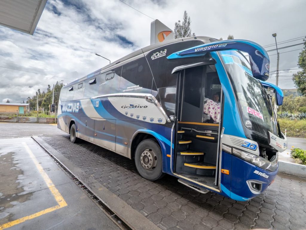 A blue and silver Amazonas bus labeled “ejecutivo” is parked at a station with the destination sign “Cuenca” displayed, representing a typical Baños to Cuenca bus option.