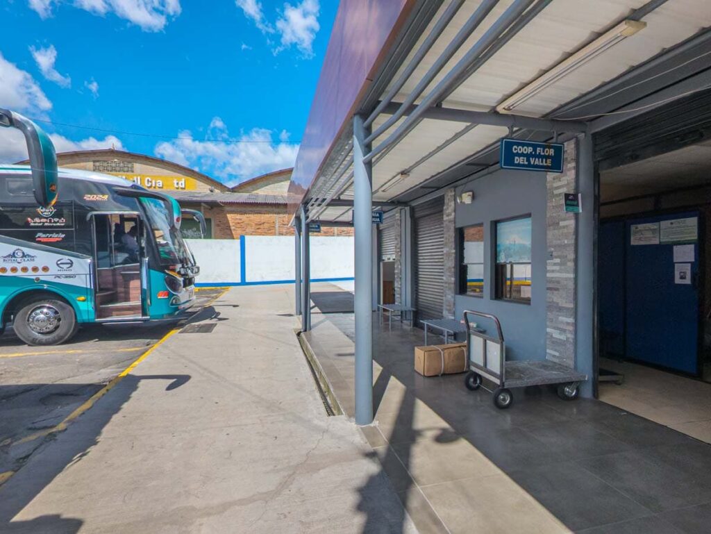 At the bus station in Quito, buses are parked in front of a sign advertising the Coop Flor del Valle ticket booth which offers direct buses from Quito to Mindo.