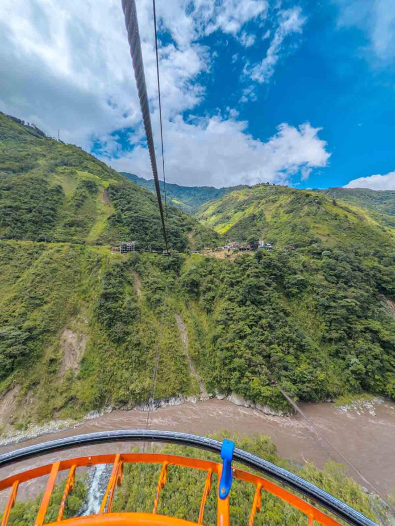 A scenic ride on a bright orange cable car crossing a river valley with lush green hills below, part of the Baños waterfall tour experience.
