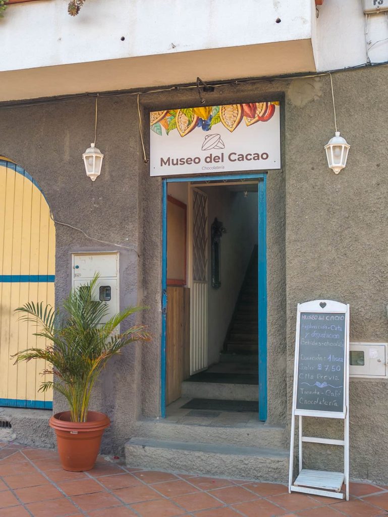 Entrance to the Museo del Cacao in Cuenca, featuring a colorful sign that says Museo del Cacao. Out front is a chalkboard listing tasting and chocolate workshop information.