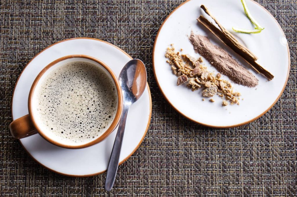 A cup of cafe de olla served on a saucer with a spoon beside a small plate of cinnamon sticks piloncillo, lime zest, and ground spices. The warm spiced Mexican coffee is a traditional drink, also enjoyed during Day of the Dead.