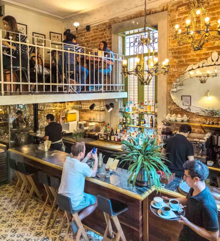 Inside Cafe Nin, a coffee shop in Mexico City, customers sit at the bar and upper balcony level while servers and baristas deliver drinks to the customers. A fancy mirror hangs on the brick wall while decorative lighting hangs from the ceiling.