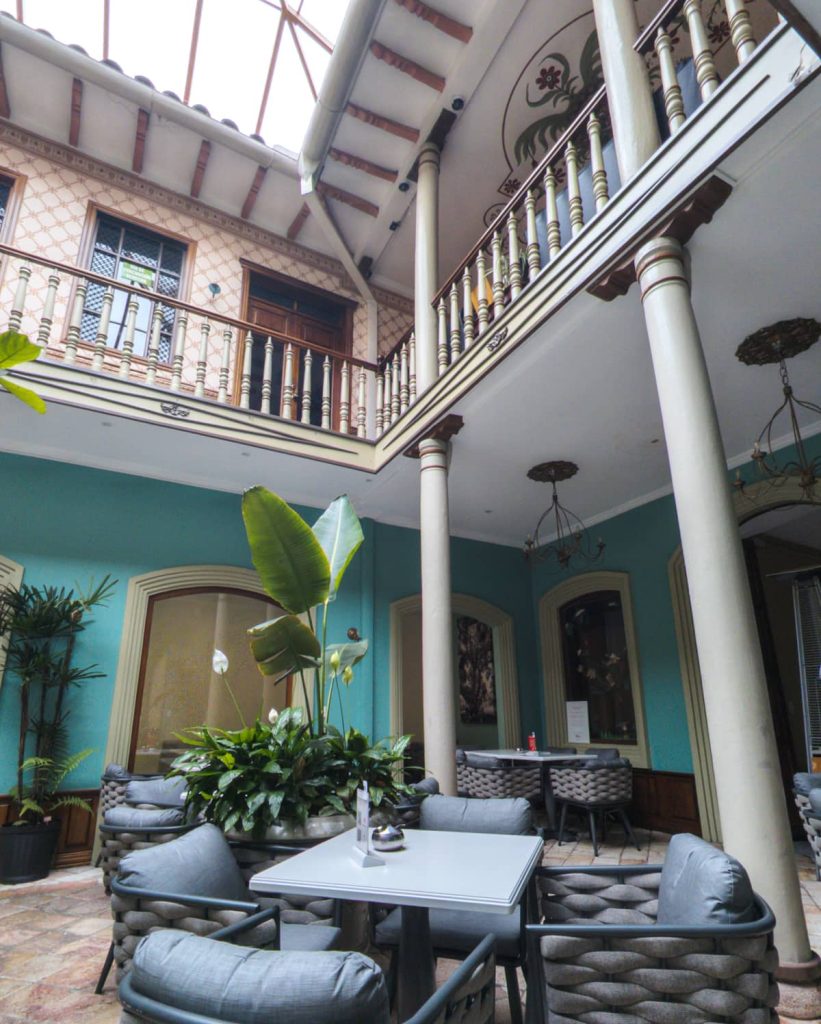 Interior courtyard of the Paccari Chocolate in Cuenca, Ecuador, featuring soft gray seating, potted tropical plants, and traditional architecture with wooden balconies and columns.