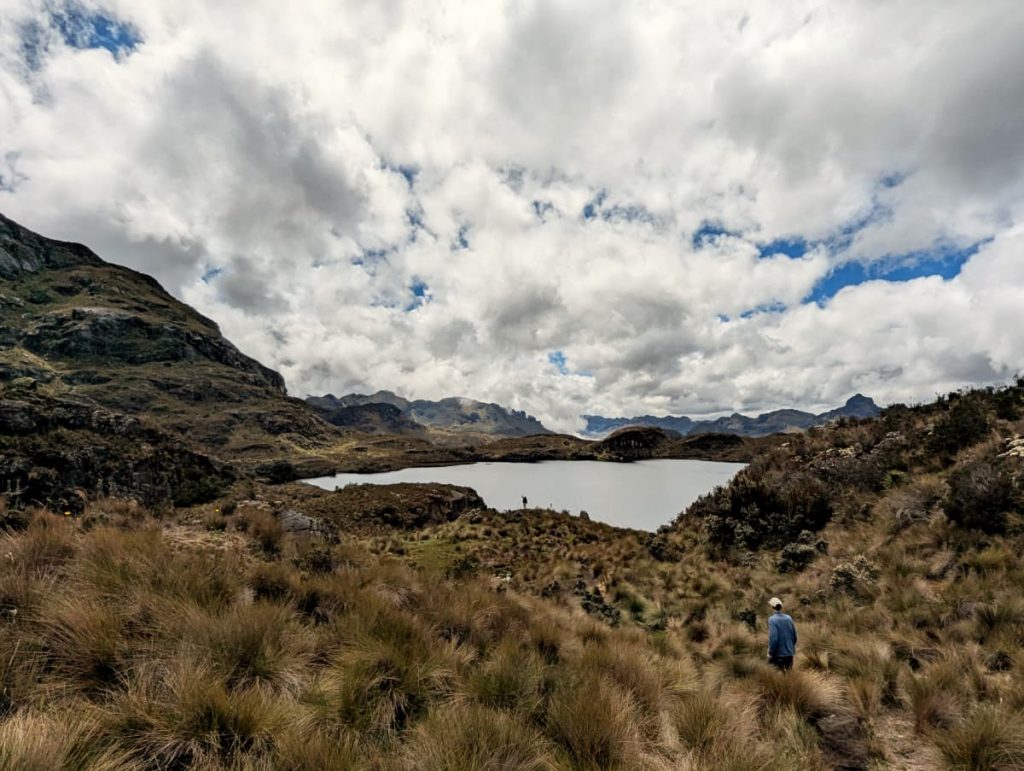 Two hikers explore the rugged grasslands of Cajas National Park, surrounded by dramatic Andean peaks and a reflective mountain lake under a sky filled with thick, textured clouds. The expansive landscape captures the park’s remote beauty, making it a stunning day tour from Cuenca.