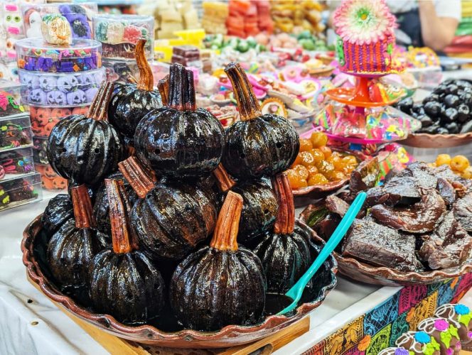 Glossy whole candied pumpkins, calabaza en tacha, stacked in a clay dish at a colorful market stall. The dark syrup coated pumpkin is a classic Day of the Dead food often placed on ofrendas as a sweet offering. In the background are plates of candied fruits and other sweet treats.