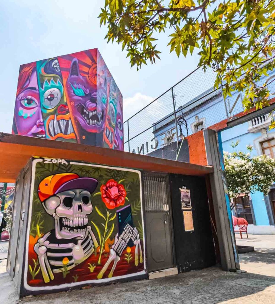 On Calle Regina, street art depicts a skeleton wearing a baseball cap and holding a cellphone. Above it, a mural of multiple monster faces with big teeth curve around the corner of the structure.