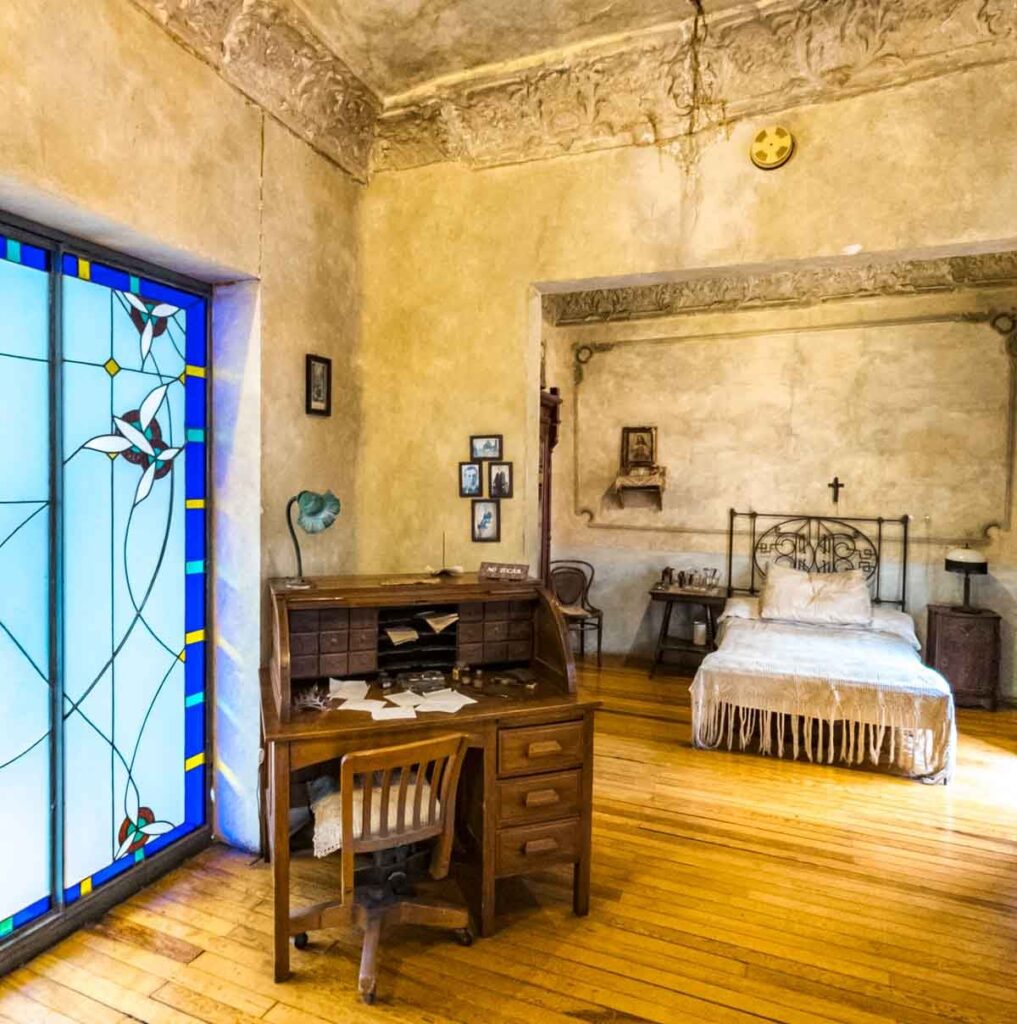A bedroom with a bed, and a wooden study table next to blue glass windows at Casa del Poeta in Mexico City.