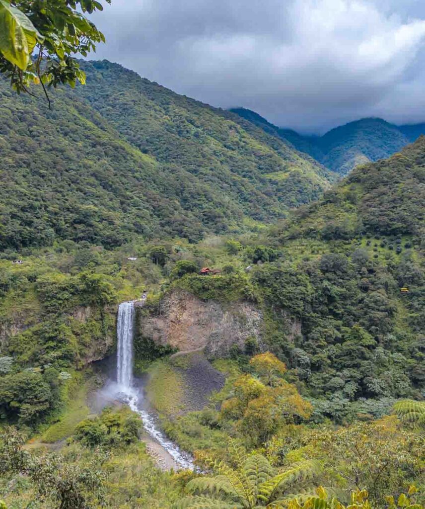 A tall, narrow waterfall cascades over a lush green cliff surrounded by dense forest in the mountains near Baños Ecuador. Mist rises from where the water hits the rocks below.
