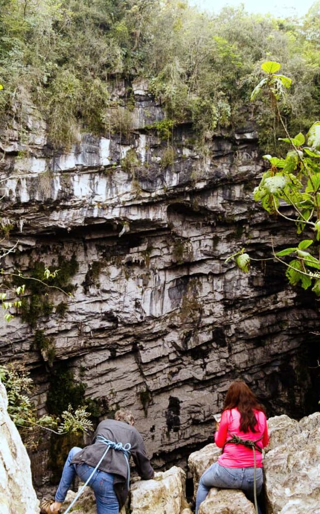 A man and a woman lean over the edge of The Cave of Swallows with climbing robes tied around their waists. In the background you can see the jagged rocky wall of the cave.