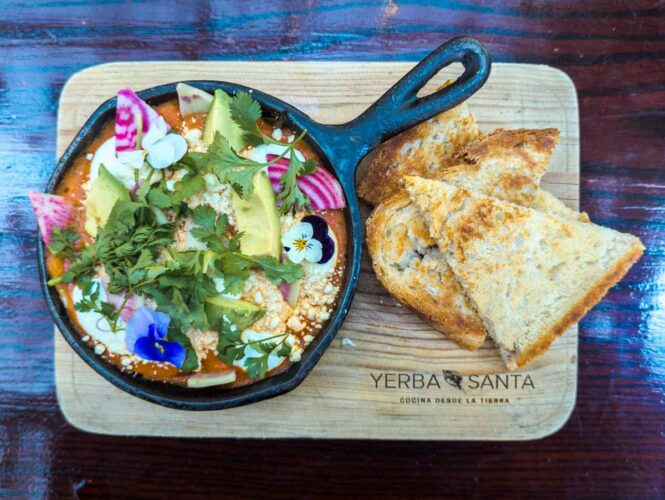 A dish of eggs baked in tomato sauce and topped with slices of avocado, radish, cilantro, and edible flowers is served in a small cast iron pan on top of a wooden cutting board with several slices of toasted bread. The cutting board is stamped with the logo of Yerba Santa, a restaurant with one of the best breakfasts in Mexico City.