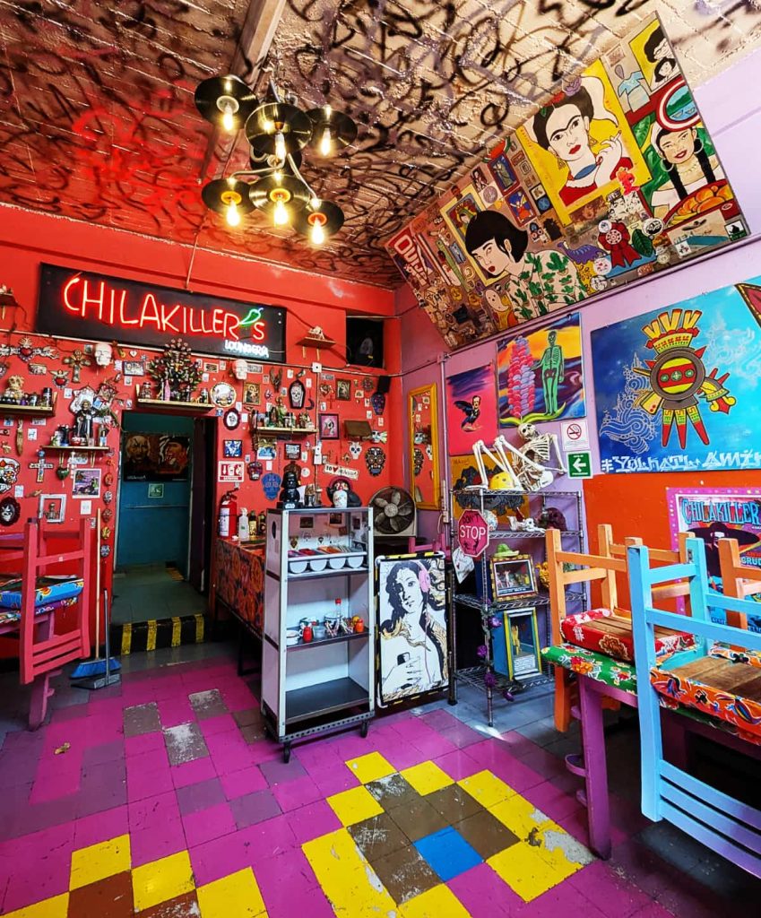 The colorful interior of Chilakiller's Loungeria in Mexico City, with red walls covered in Mexican folk art, a neon sign of the restaurant name with a jalapeno for the apostrophe, and a chandelier made from small vinyl records