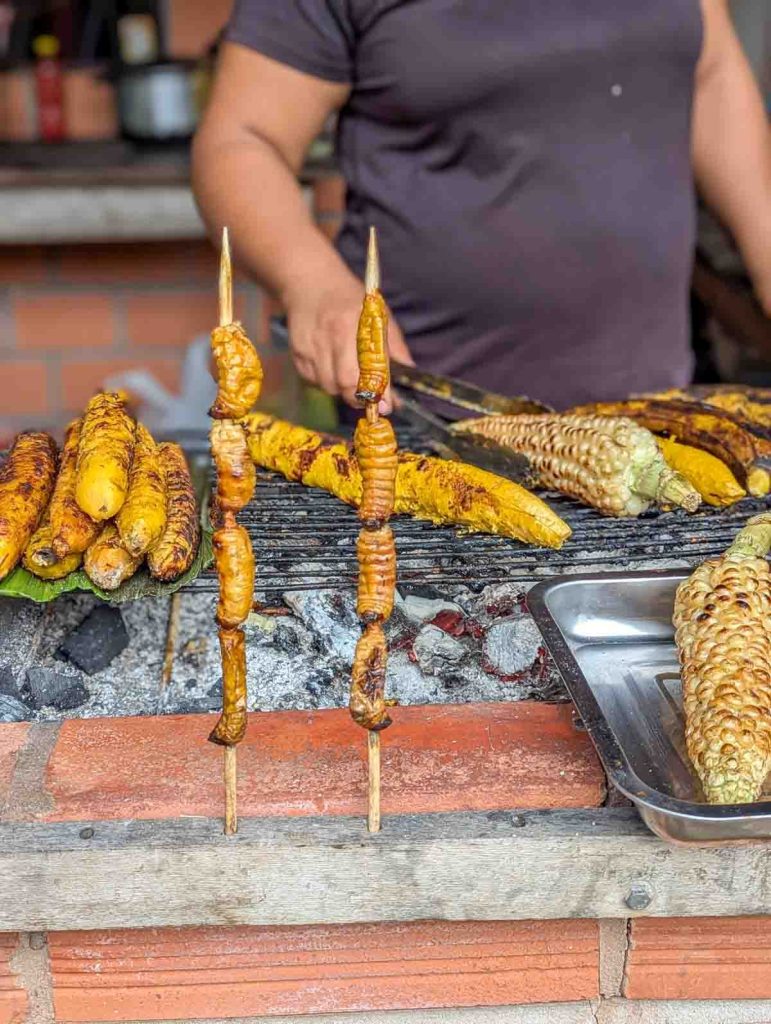 Skewers of grilled chontacuros, a unusual street food in Ecuador, that have been roasted over hot coals. In the background a woman flips corn on the grill next to a pile of grilled plantains.