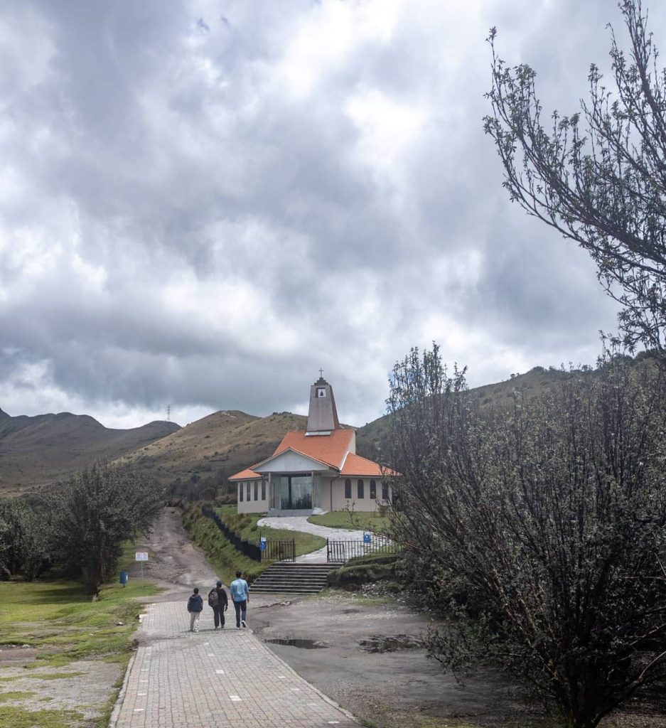 Three people walk toward a small chapel with a red roof nestled in the hills, near the top station of the Teleferico de Quito.