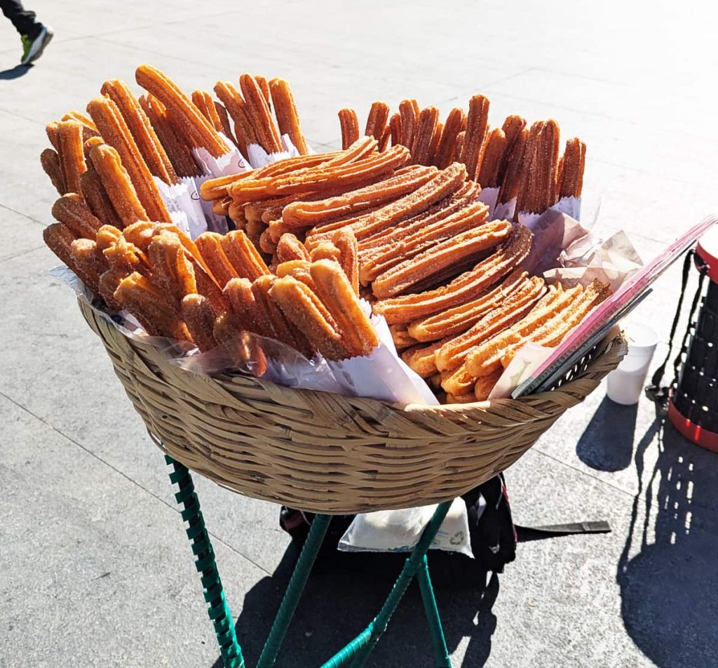 Golden, sugar-dusted churros stacked in a large wicker basket on a street in Mexico City. Around the edge are white paper bags with 4-5 churros each and in the middle is a large stack of churros for individual sale.