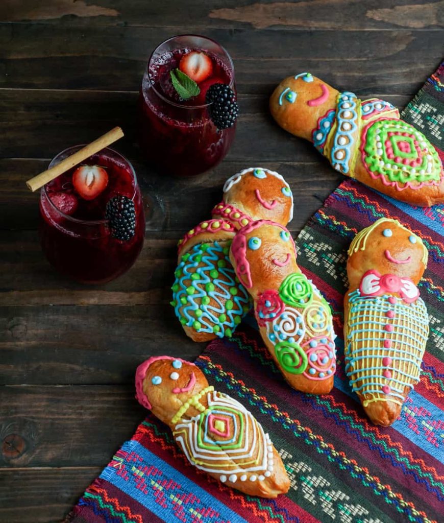 Colorful guaguas de pan decorated with bright icing lay on a traditional woven cloth beside two glasses of colada morada, a deep purple Ecuadorian drink garnished with strawberries, blackberries, and a cinnamon stick.