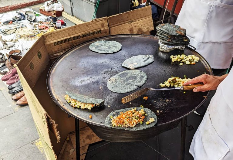 A Mexico City street food vendor uses a spatula to cook blue corn tlacoyos and quesadillas on a large round black cooking surface called a comal. At the street stand next door a white sheet with shoes, belts, and other used items are for sale.