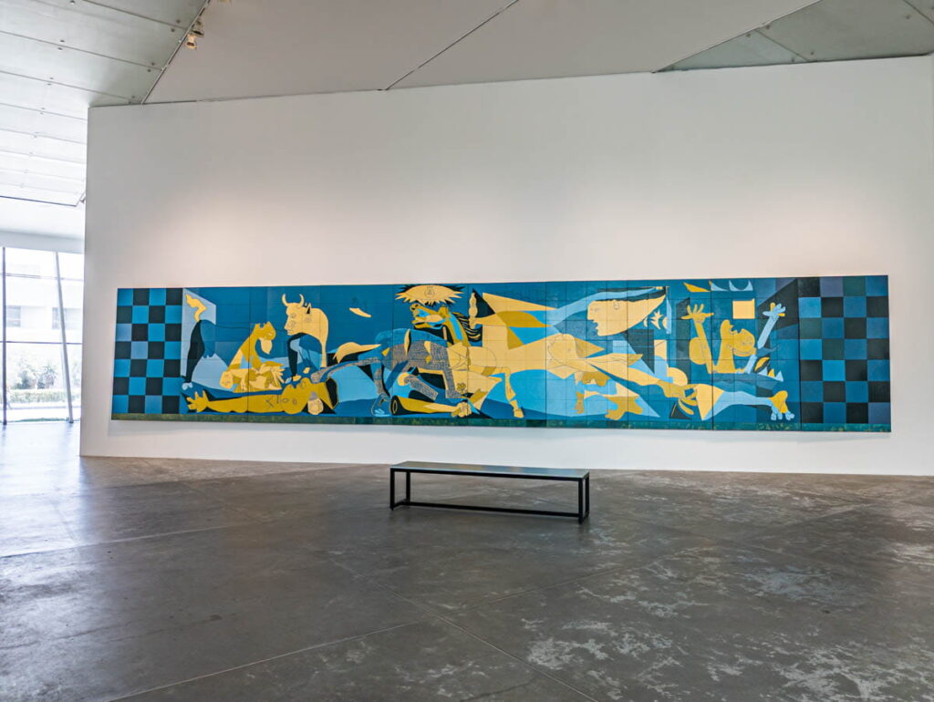 A long blue and yellow artwork in a white room at the Contemporary Art Museum in Mexico City. In front of the contemporary artwork is a simple black bench.