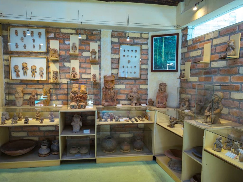 Display of pre-Columbian ceramic figurines and artifacts inside a Cuenca museum, arranged against exposed brick walls with framed collections and spot lighting.
