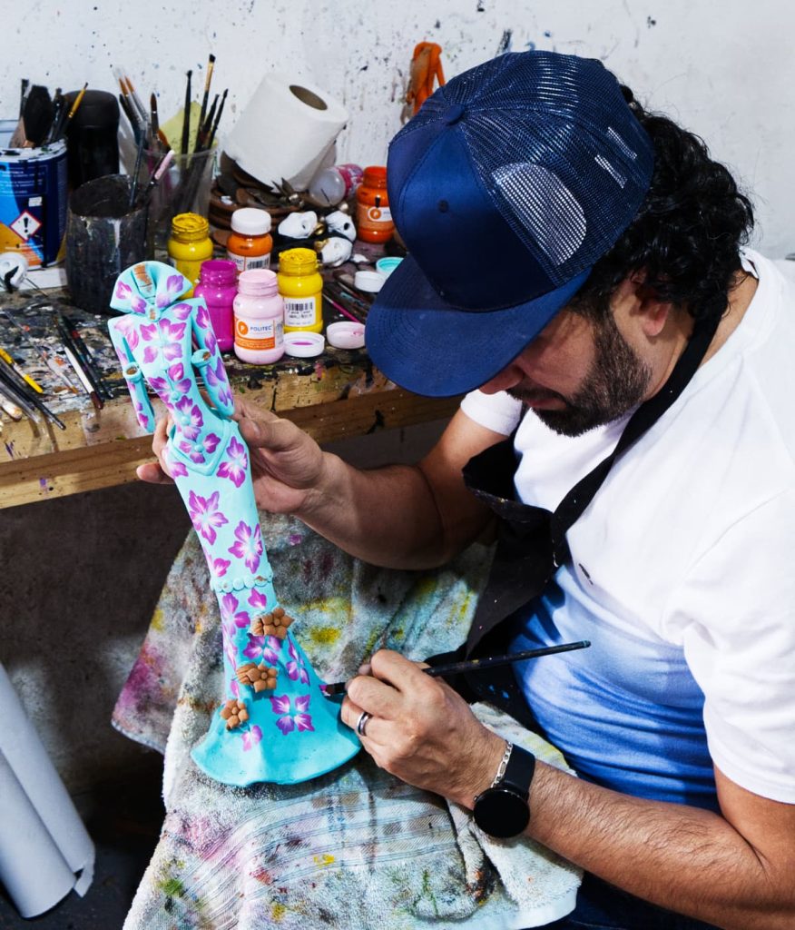 Artist hand-painting a turquoise La Catrina figurine with pink flowers at a workshop in a Capula Michoacán surrounded by brushes and paint jars.