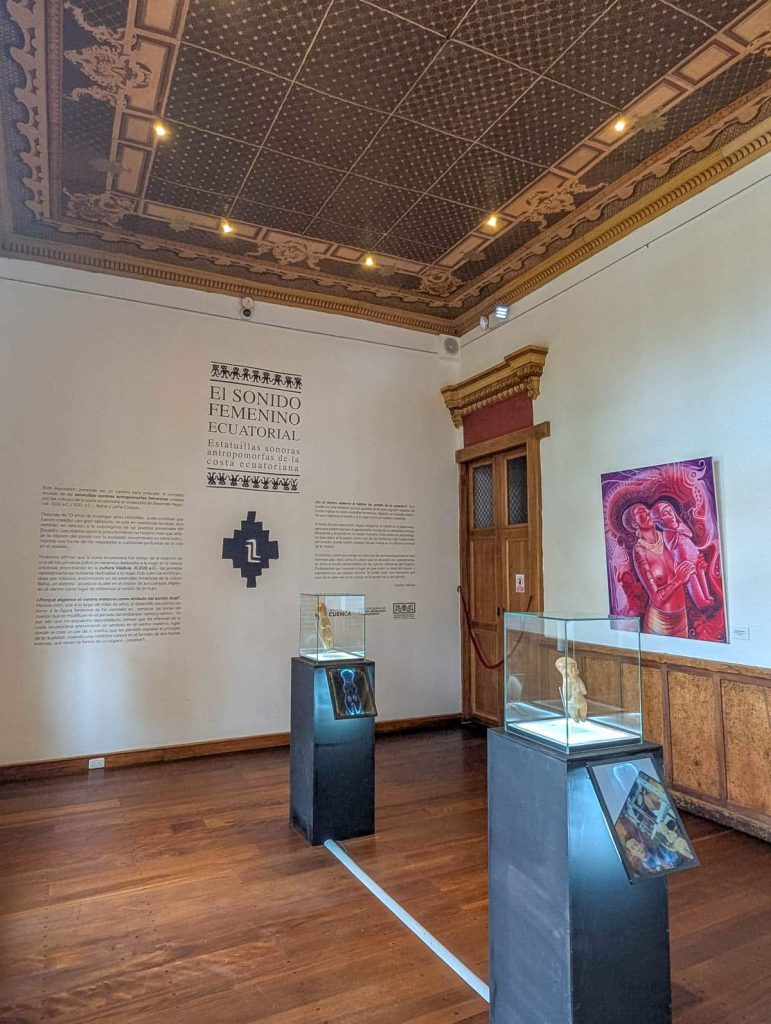 Exhibit titled "El Sonido Femenino Ecuatorial" inside a Cuenca museum, featuring Ecuadorian anthropomorphic sculptures and a richly decorated ceiling.