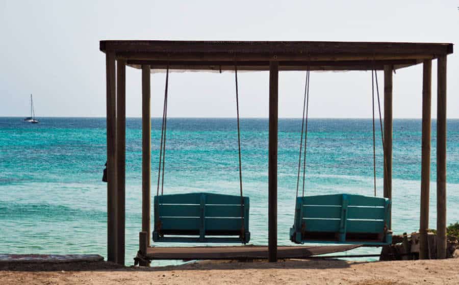 Wooden porch swings overlook the blue ocean at El Hamaquero as a sailboat passes by in the distance.