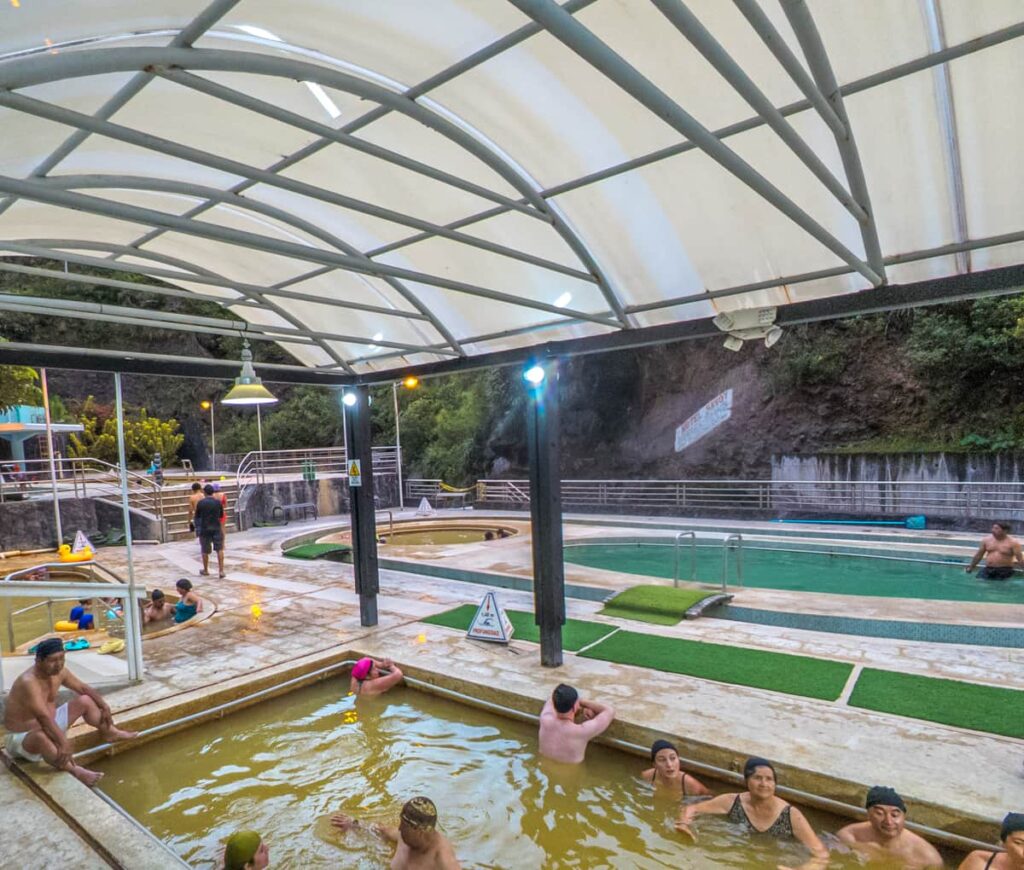 People relax in multiple thermal pools at a covered outdoor hot spring in Baños Ecuador, with mineral-rich yellow and green waters. The pools are surrounded by mountain rock and greenery.