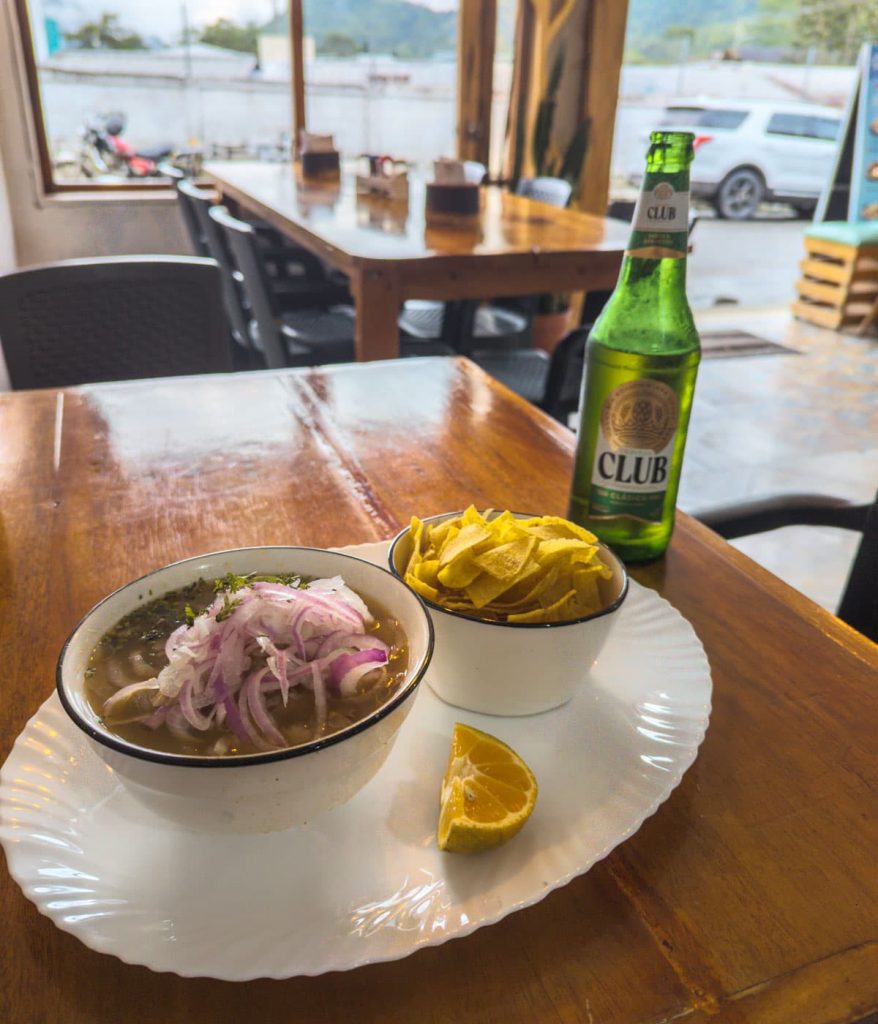 A bowl of encebollado, one of the most traditional Ecuadorian foods, topped with sliced red onions and cilantro, served with a bowl of cripsy plantain chips and lime. Sitting on the wooden table is a green bottle of Club beer, a favorite alcoholic drink in Ecuador.