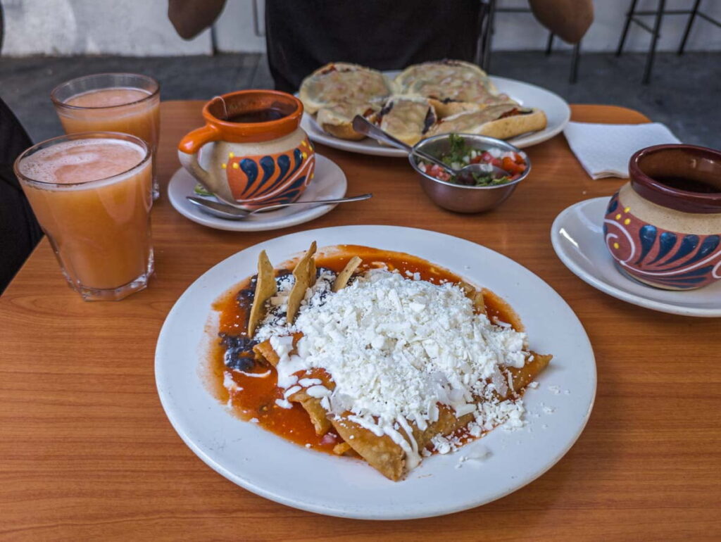For a cheap breakfast in Mexico City, enchiladas are bathed in a red sauce and topped with cheese. Also on the table are cups of coffee, tea, and juice.